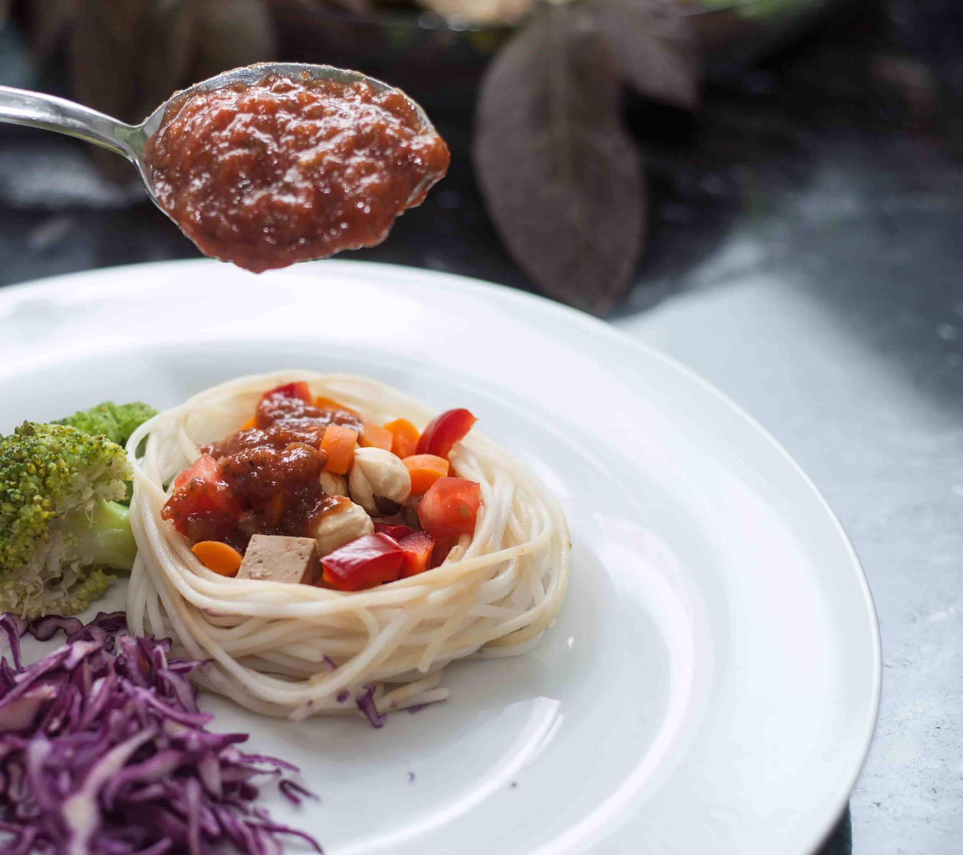 Un plato con un nido de pasta cubierto con salsa de tomate y verduras, con una cuchara suspendida. Brócoli y col lombarda como acompañamiento.