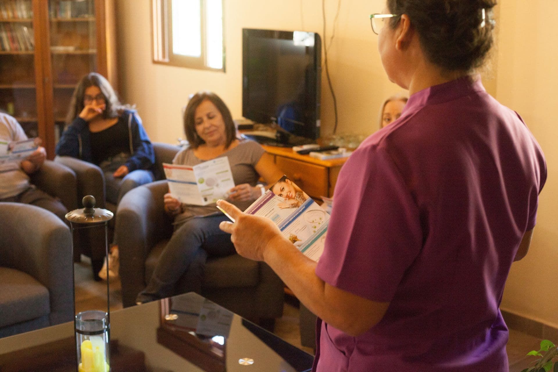 Persona con una blusa morada se presenta ante un pequeño grupo sentado en una sala de estar y señala un documento.