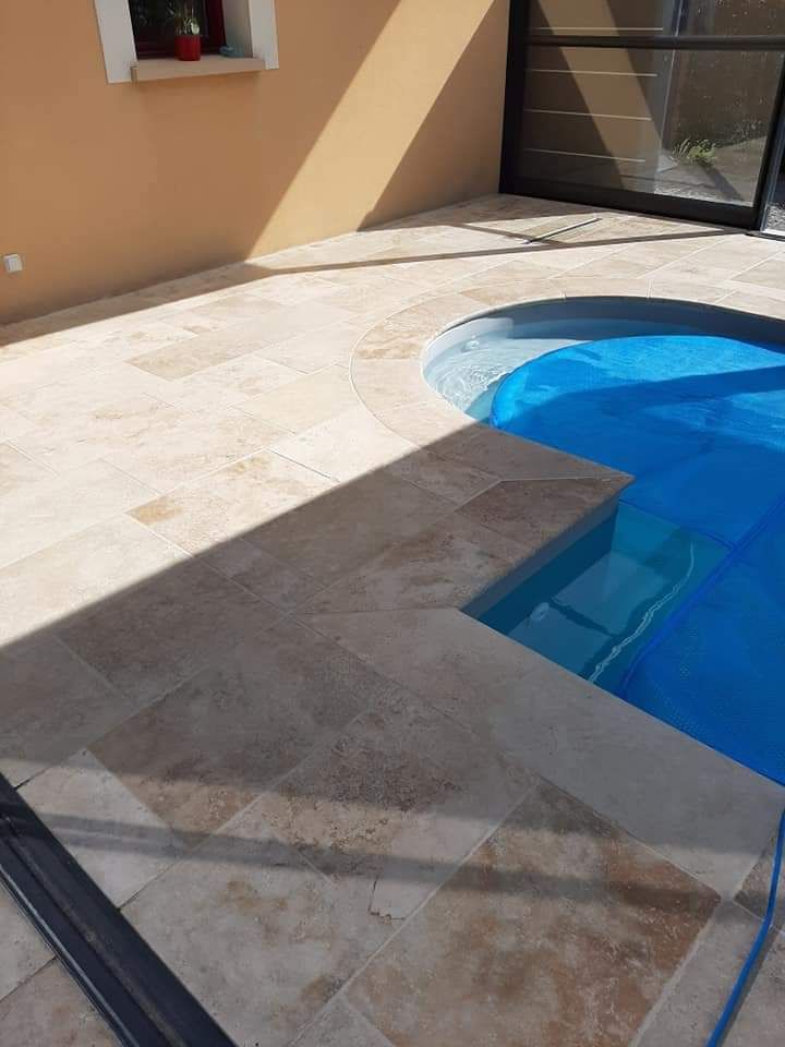 Piscine recouverte de bleu, entourée d'une terrasse en pierre claire. Mur marron et porte sombre en arrière-plan.