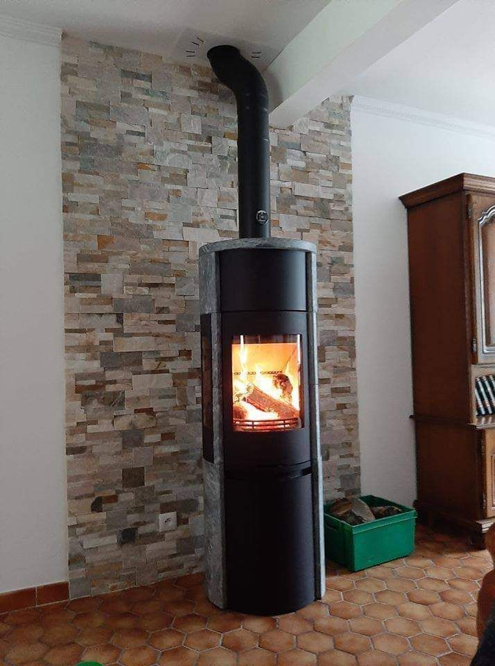 Poêle à bois avec feu allumé contre un mur carrelé. Conduit de cheminée noir, bois de chauffage dans une caisse verte.