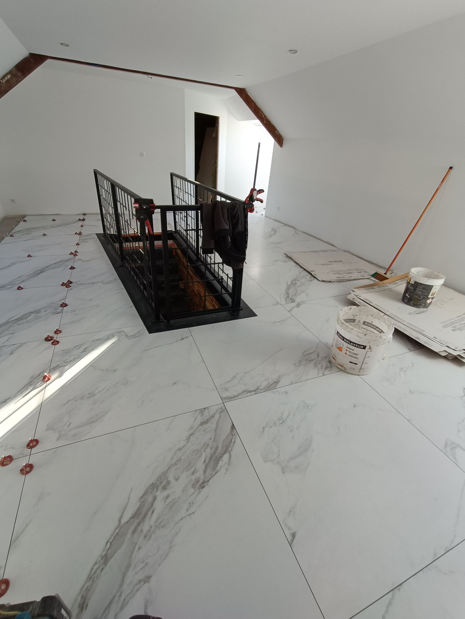 Vue intérieure d'une pièce en construction avec des carreaux blancs imitation marbre. Une rambarde noire entoure la cage d'escalier.
