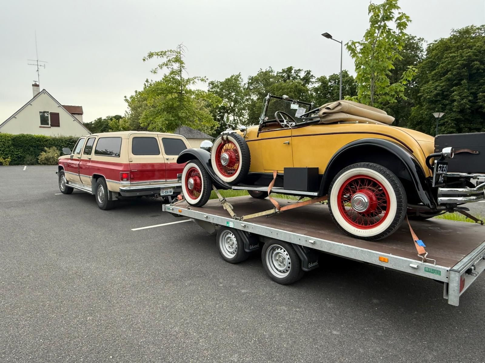 Une voiture décapotable jaune classique sur une remorque, tractée par un SUV rouge et beige sur un parking goudronné.