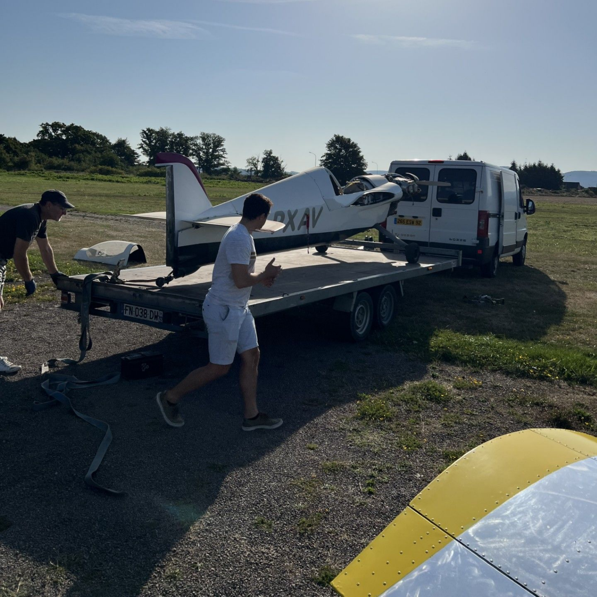 Deux hommes chargent un petit avion sur une remorque. L'avion est blanc avec des touches de rouge, sur un terrain de gravier.