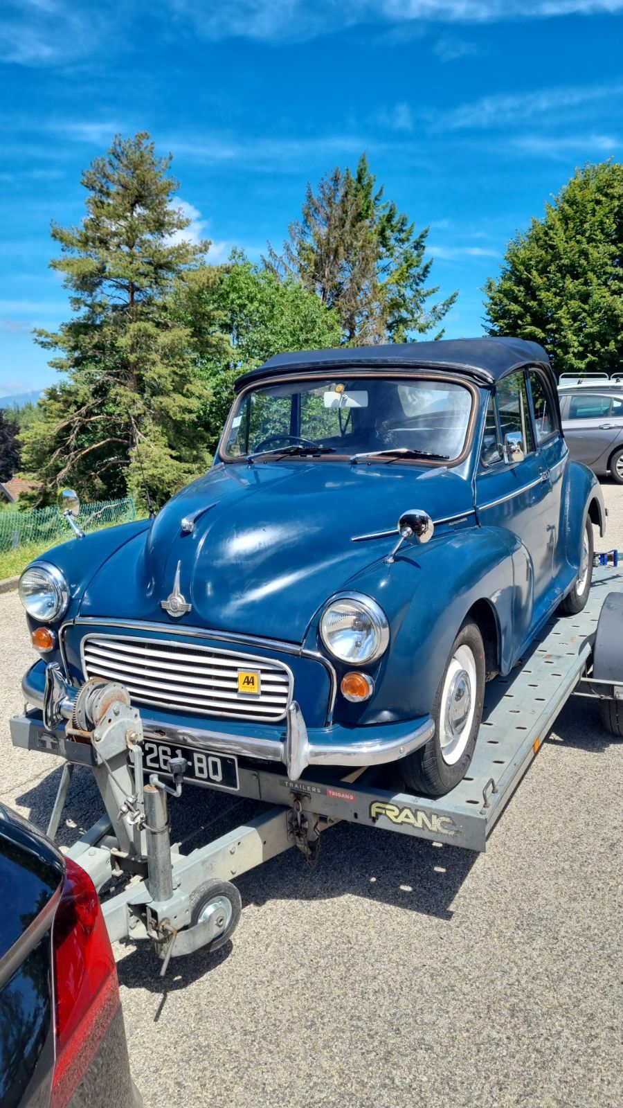 Une Morris Minor décapotable bleue vintage sur une remorque, garée à l'extérieur par une journée ensoleillée.