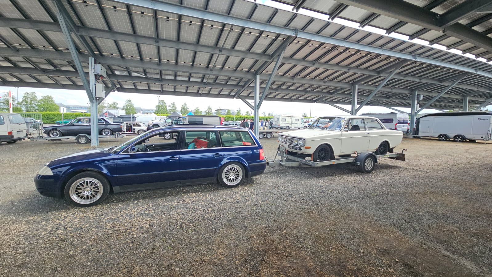 Un break bleu tracte une voiture blanche sur une remorque sous un parking couvert.
