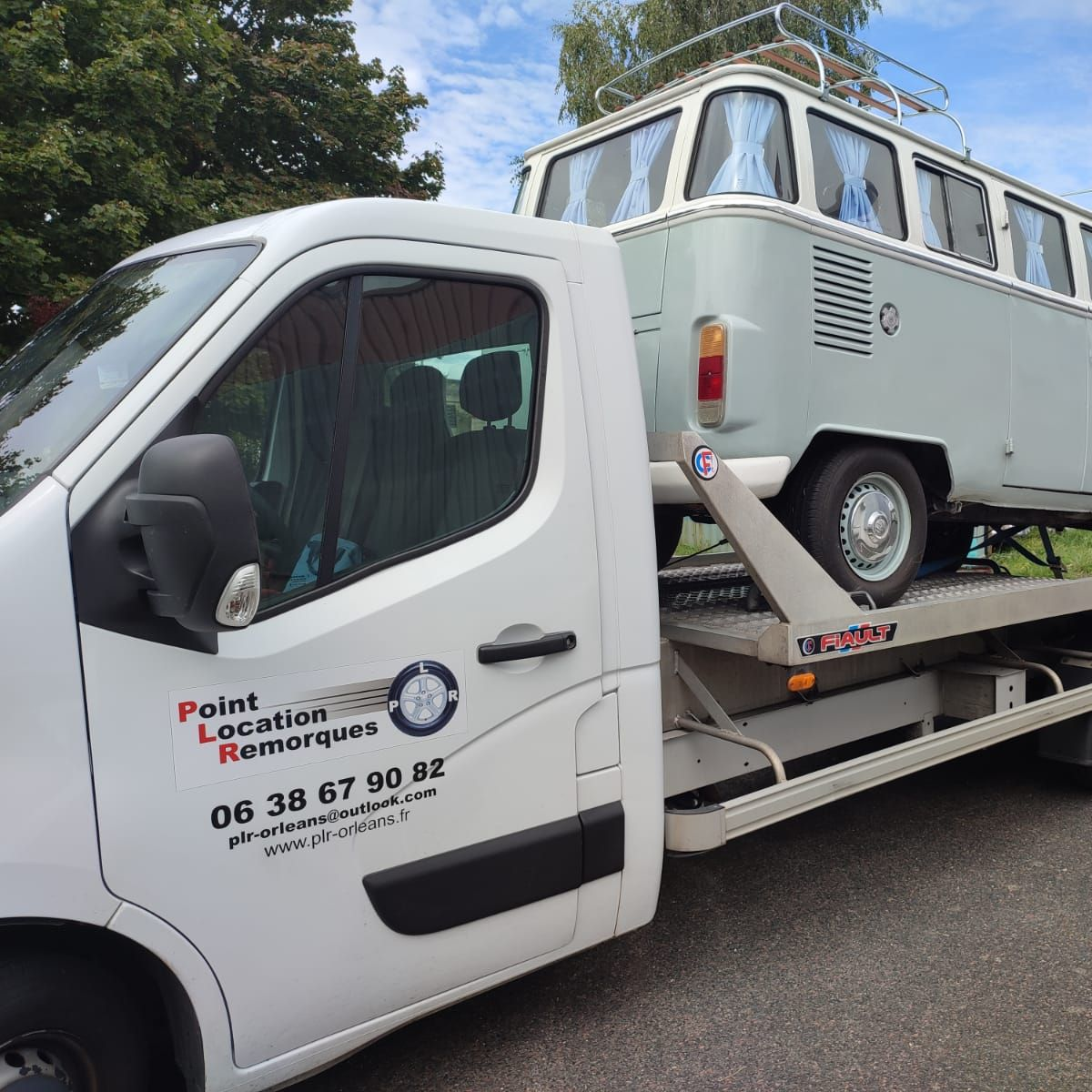 Un camion de dépannage blanc à plateau transportant un camping-car Volkswagen bleu clair vintage sur une route de campagne.