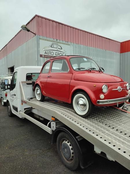 Une Fiat 500 rouge vintage sur une dépanneuse blanche. Garée devant un garage à la façade rouge et grise.