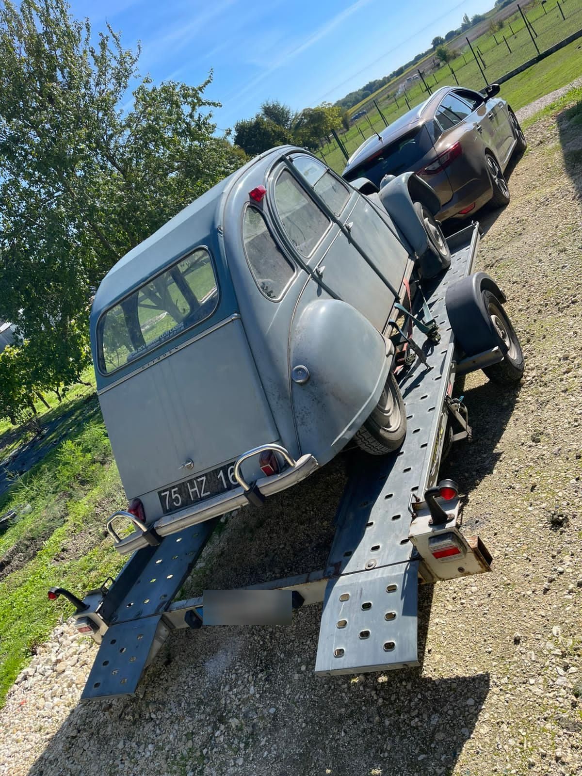 Une Citroën 2 CV grise sur une remorque, garée sur une pelouse, sous un ciel bleu.