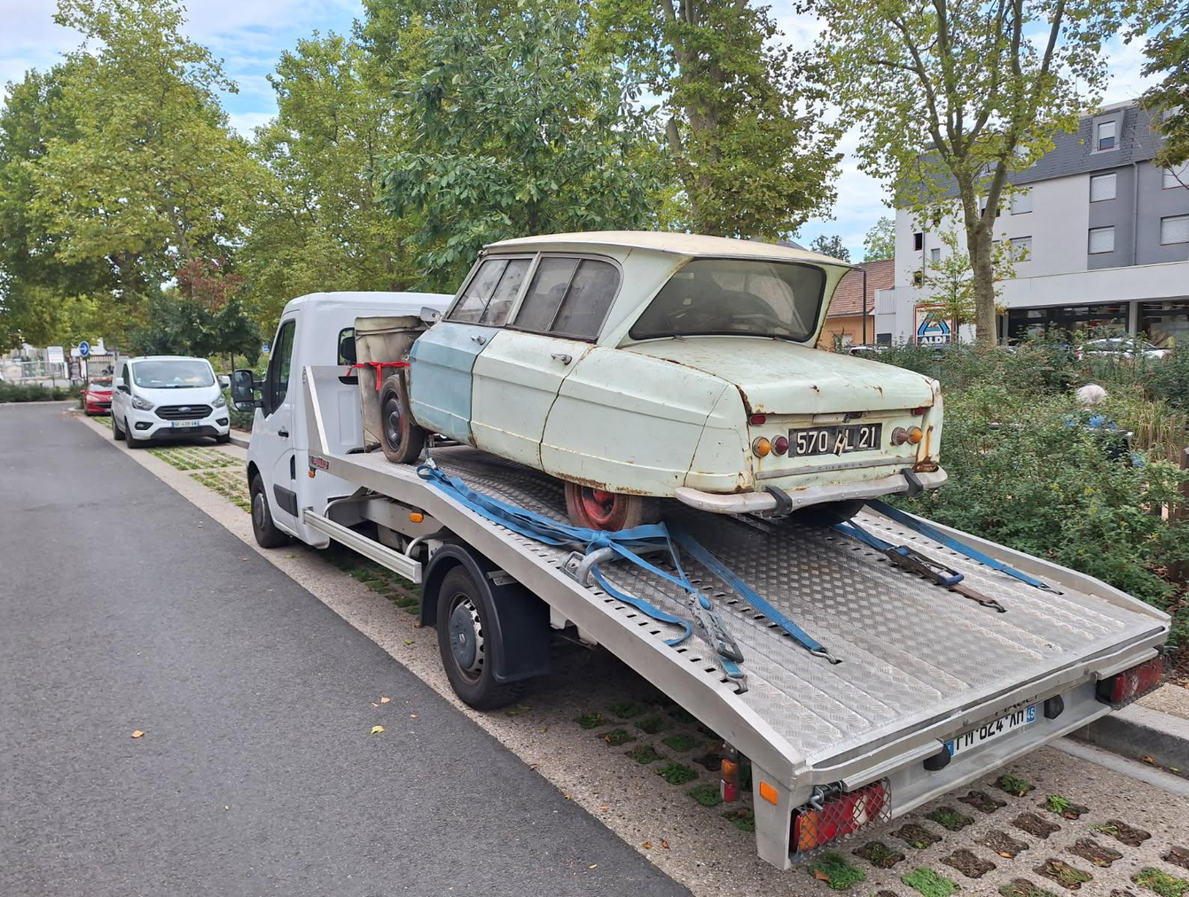 Une dépanneuse blanche transporte sur son plateau une voiture classique bicolore usée par le temps, sur un parking.