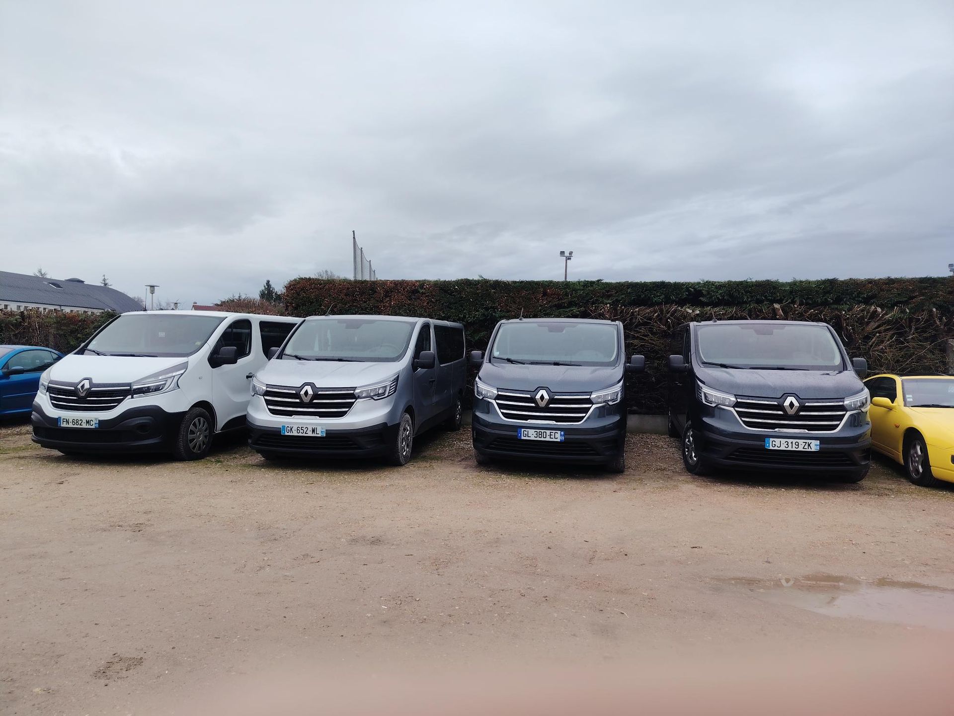 Quatre fourgonnettes Renault Trafic garées en rang sur un terrain gravillonné, à côté d'une voiture jaune, avec une haie en arrière-plan.