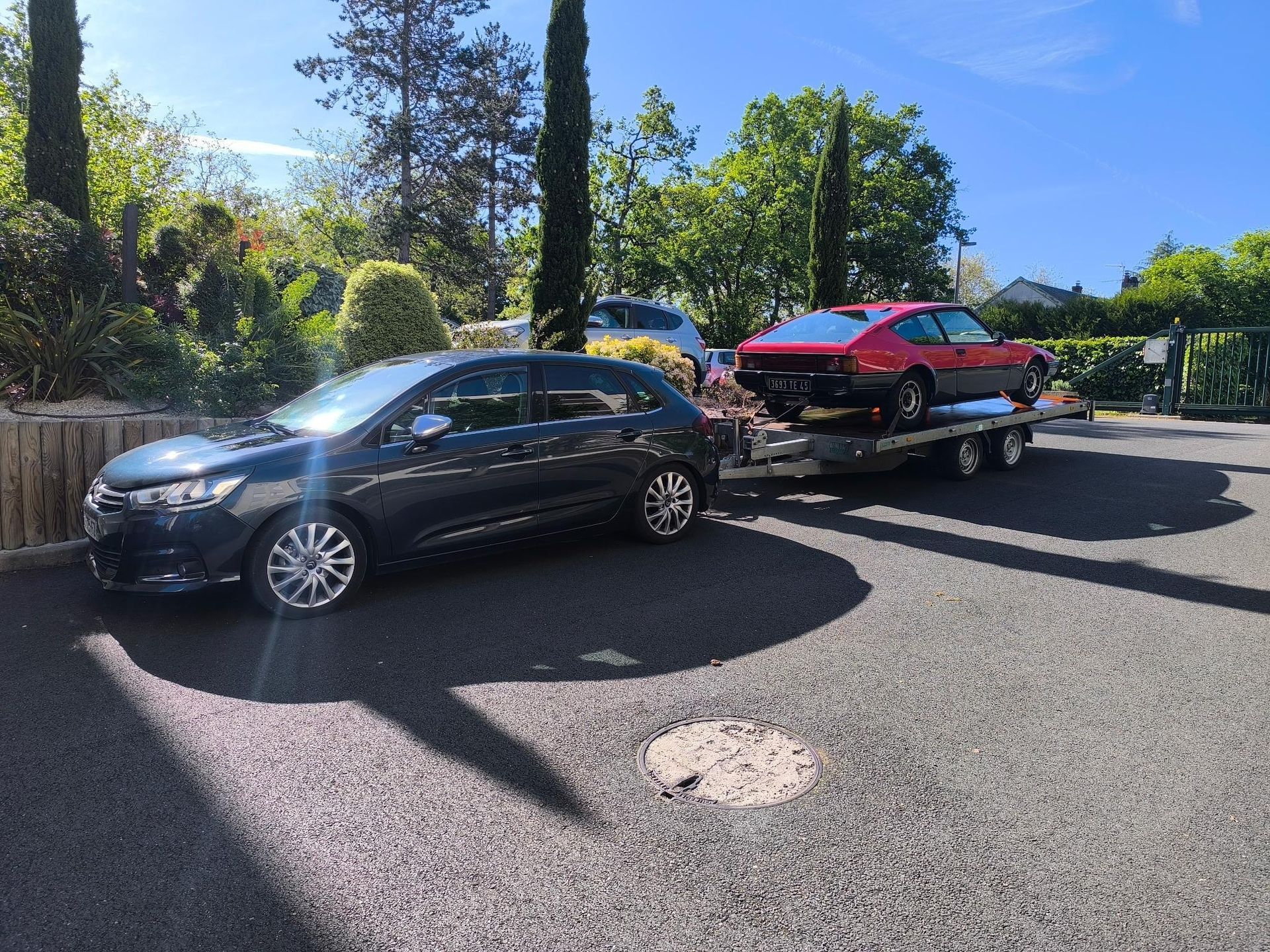 Une berline noire tracte une voiture rouge sur une remorque dans une rue résidentielle ensoleillée.