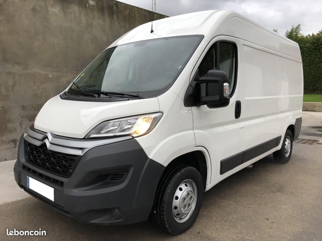 Une fourgonnette Citroën Jumper blanche est garée sur un terrain goudronné, avec un mur en béton en arrière-plan.