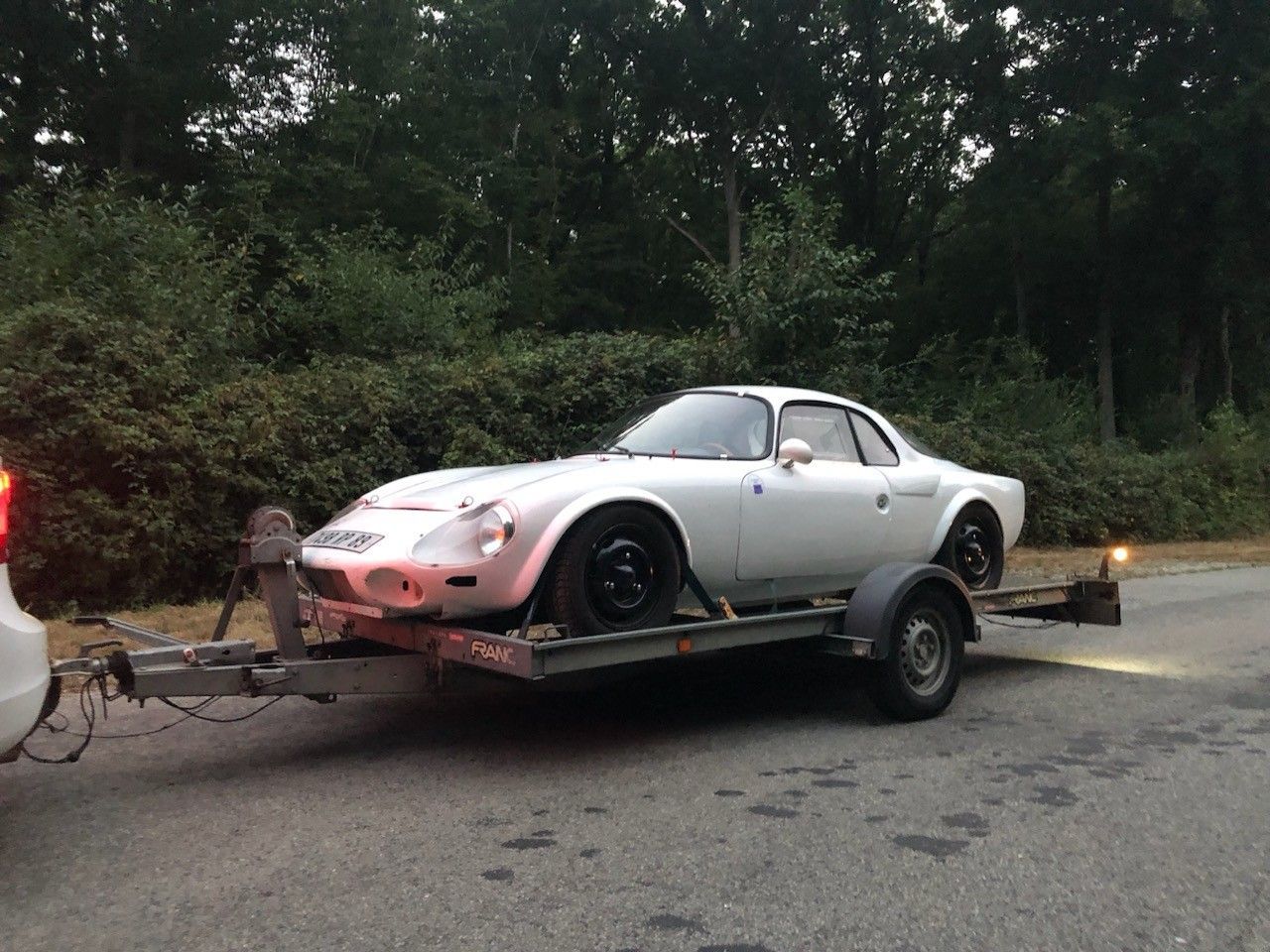 Voiture de sport blanche sur une remorque, garée sur une route avec des arbres en arrière-plan.