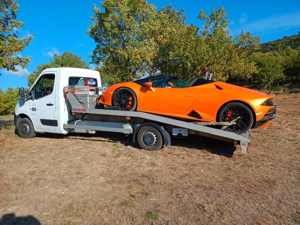 Lamborghini décapotable orange sur une dépanneuse, en extérieur avec des arbres et un ciel bleu.