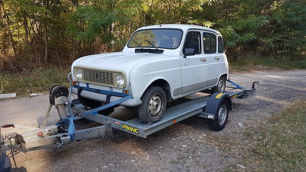 Une Renault 4 blanche sur une remorque, garée sur une surface gravillonnée à côté d'herbe et d'arbres.