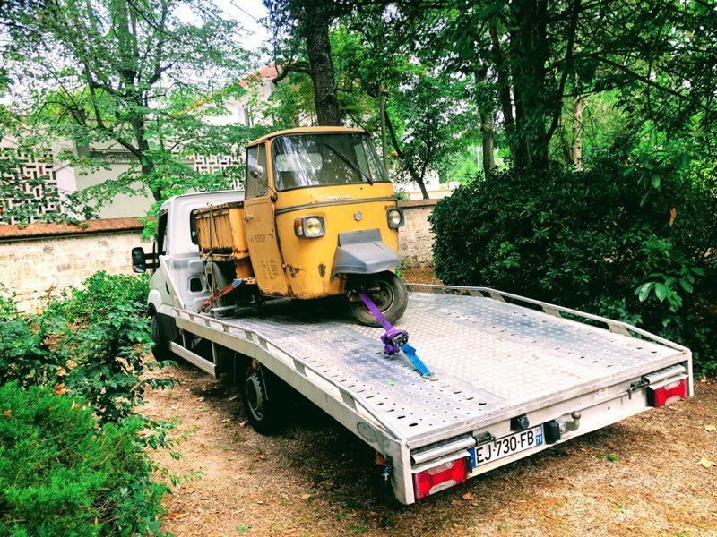 Un véhicule jaune à trois roues est chargé sur une dépanneuse blanche à plateau, à l'extérieur, près d'arbres et de buissons.