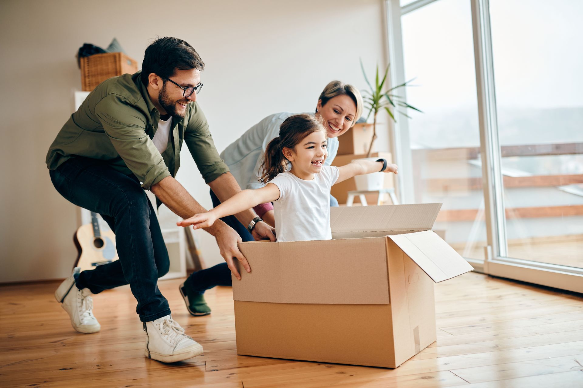 Un enfant souriant est assis dans une boîte en carton, les bras tendus, tandis que deux adultes poussent la boîte sur un parquet.