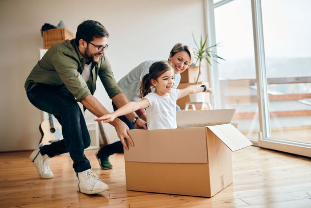 Un enfant souriant est assis dans une boîte en carton, les bras tendus, tandis que deux adultes poussent la boîte sur un parquet.