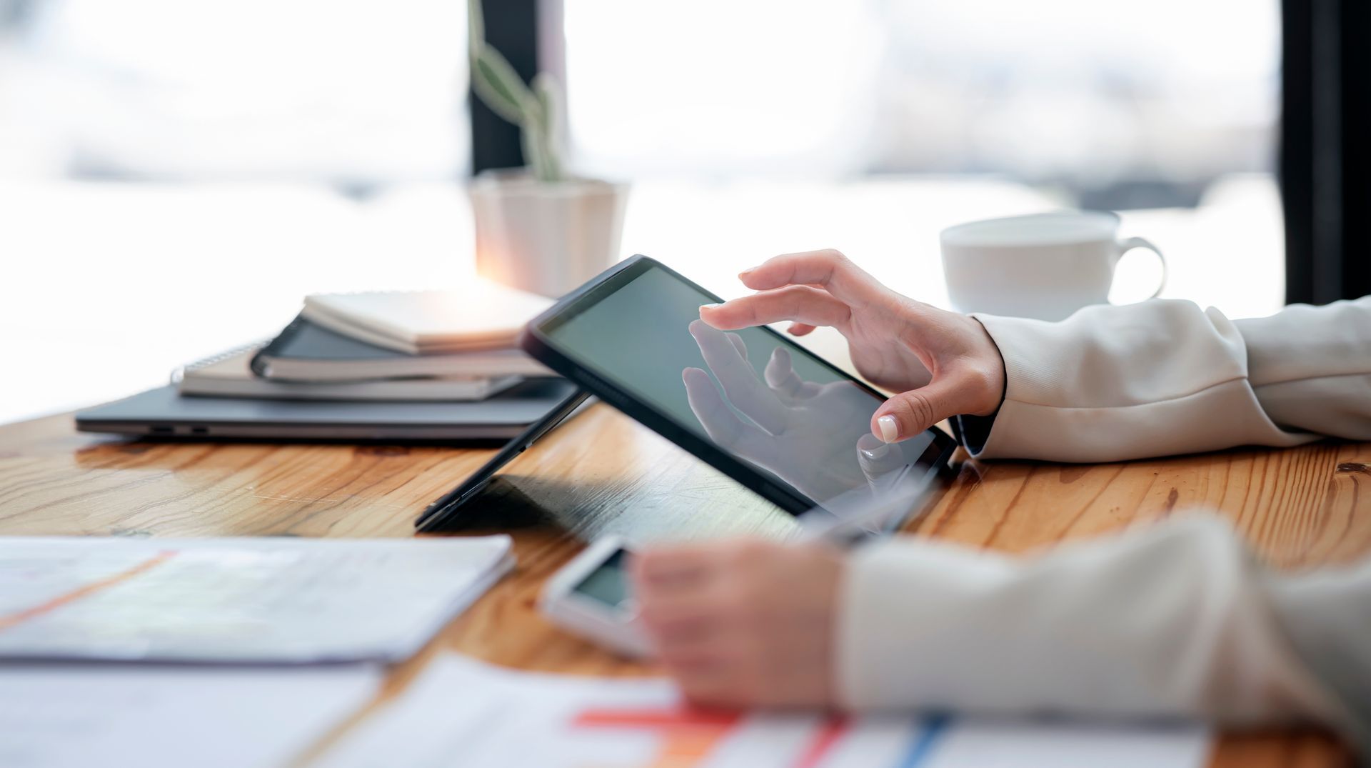 Une personne utilise une tablette sur un bureau en bois, avec des papiers, un ordinateur portable et une tasse de café à proximité.