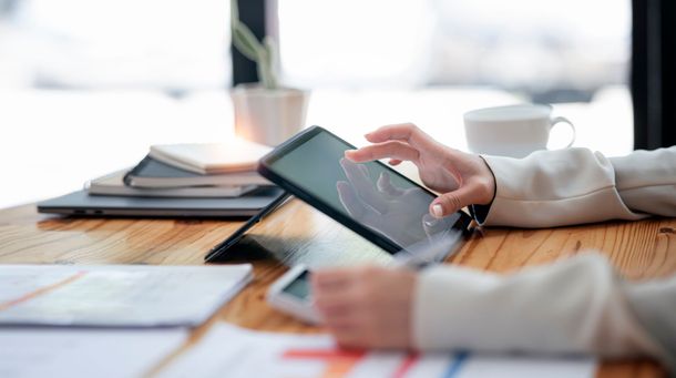 Une personne utilise une tablette sur un bureau en bois, avec des papiers, un ordinateur portable et une tasse de café à proximité.