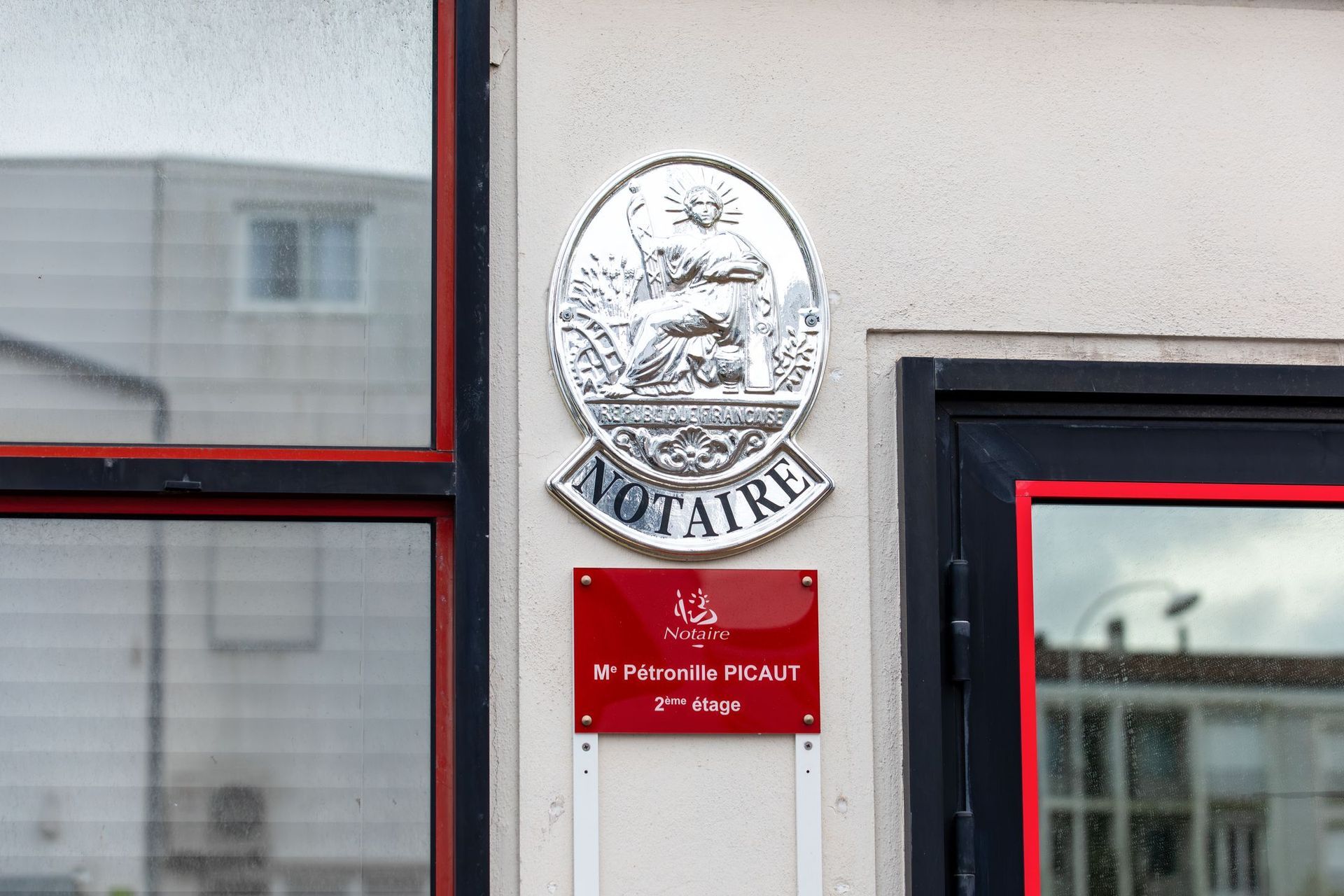 Une plaque argentée Notaire au-dessus d'une enseigne rouge pour indiquer le bureau de Mᵉ Pétronille PICAUT situé au deuxième étage d'un immeuble.