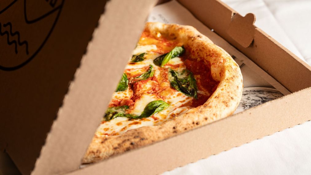 Part de pizza dans une boîte en carton avec du fromage fondu, du basilic et de la sauce tomate.