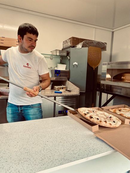Un homme en chemise blanche prépare des pizzas dans une cuisine, avec des pizzas sur le comptoir et un four en arrière-plan.