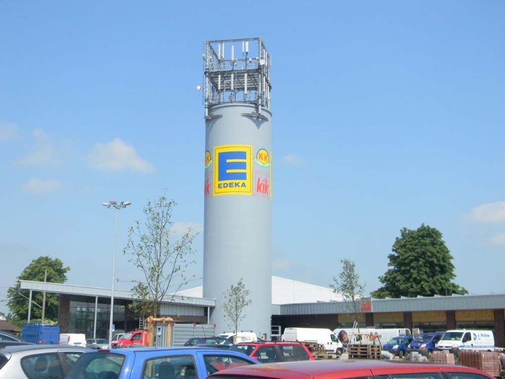 Ein hoher grauer Turm mit dem EDEKA-Logo auf einem Parkplatz an einem sonnigen Tag.