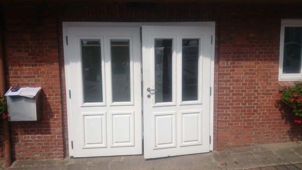 Weiße Doppeltüren mit Glaseinsätzen in einem roten Backsteingebäude; ein Briefkasten und ein Fenster sind sichtbar.