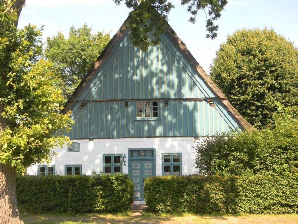 Ein weißes Haus mit blaugrüner Fassade und Strohdach, umgeben von grünen Büschen und Bäumen.