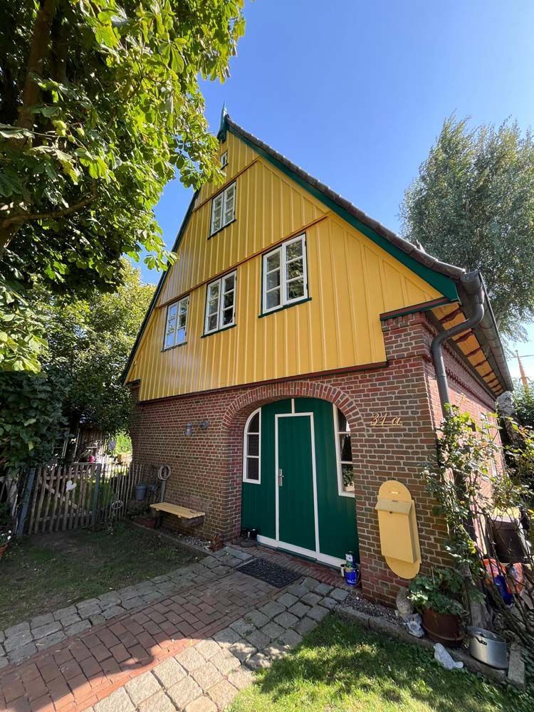 Gelbes und grünes Haus mit Ziegelsockel und gewölbtem Eingang, im Garten gelegen.