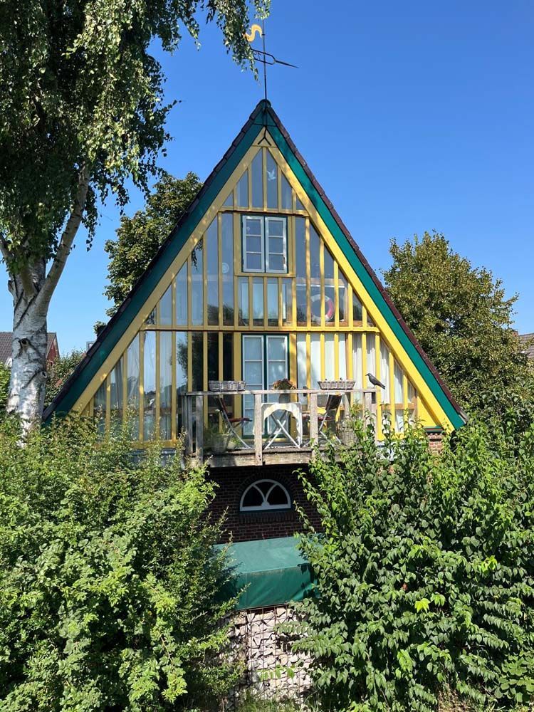 A-förmiges Haus mit gelben Zierleisten und Glasfront, Balkon, umgeben von grünem Laub unter blauem Himmel.