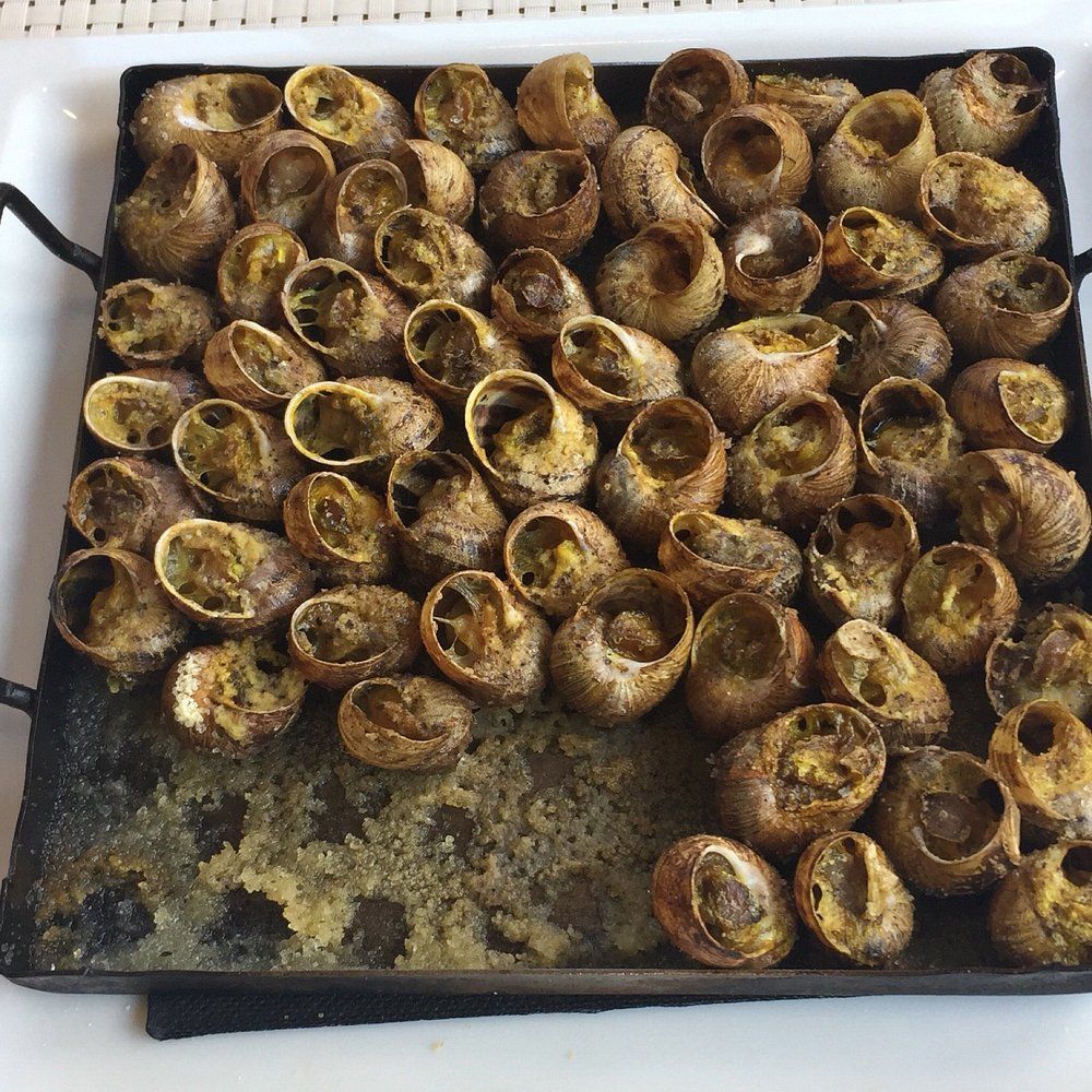 Caracoles cocidos en concha sobre una fuente de metal negro, presentados sobre una superficie blanca.