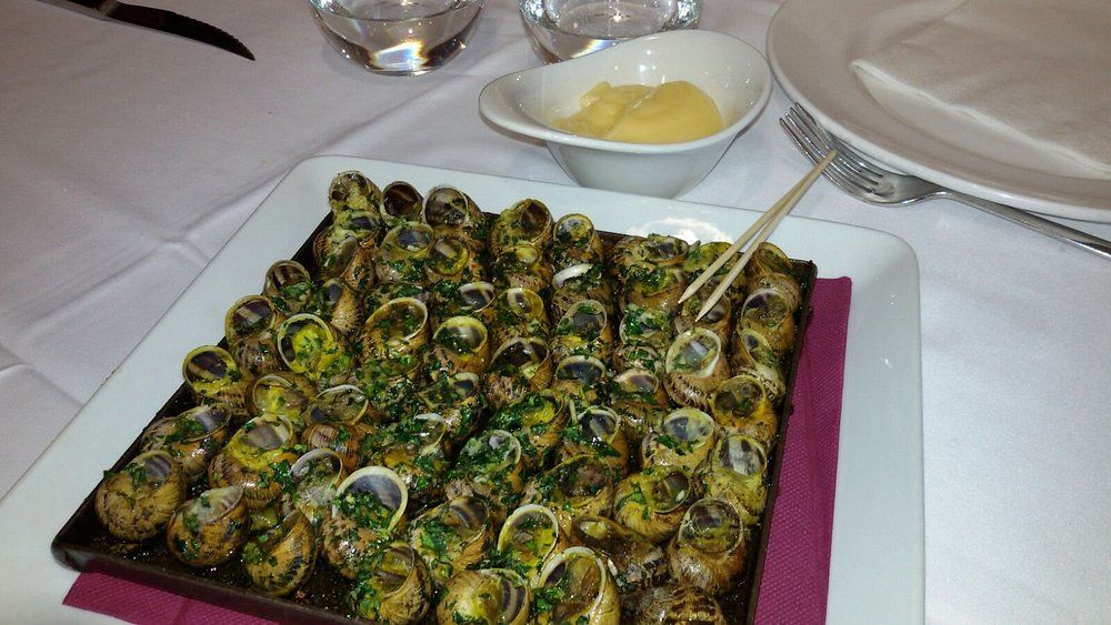 Caracoles en plato cuadrado, servidos sobre mantel blanco con mantequilla y un palillo.