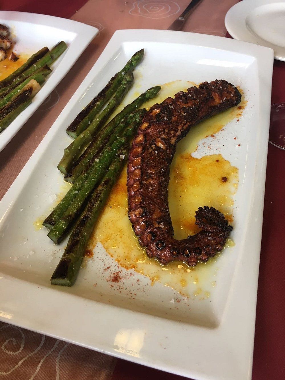 Tentáculo de pulpo a la parrilla con espárragos en un plato blanco, rociado con aceite y especias.