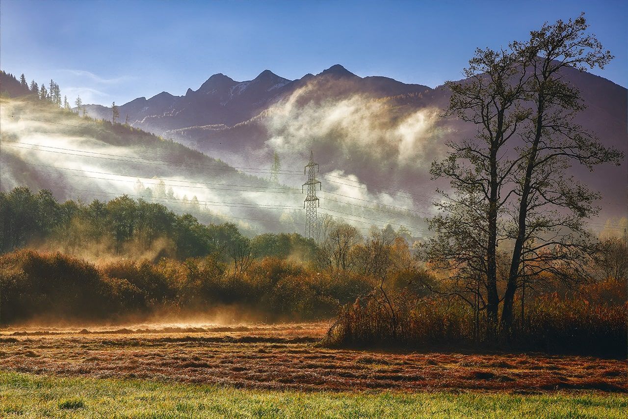 the sun is shining through the clouds over a field with trees and mountains in the background