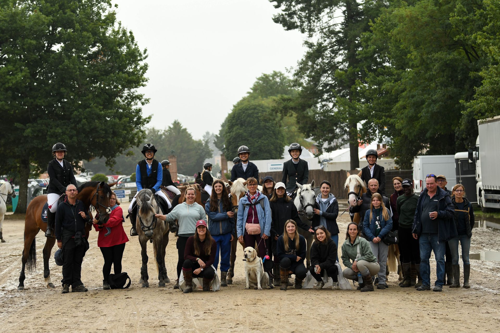 Grand groupe posant en extérieur sur un chemin de gravier, avec des chevaux et des arbres en arrière-plan.