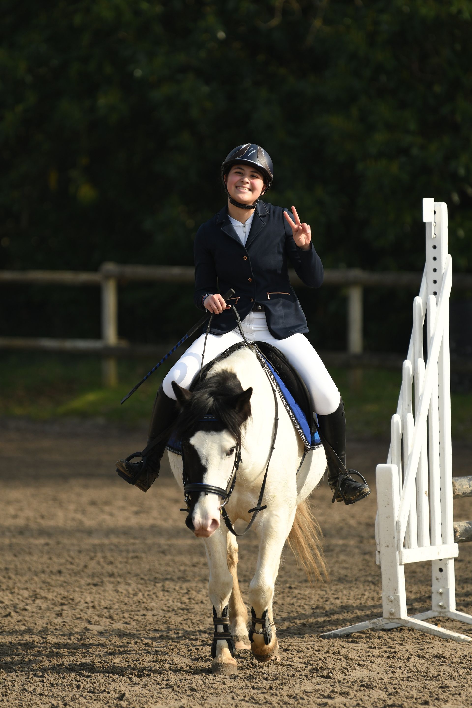 Cavalier sur un cheval noir et blanc dans un manège extérieur, souriant et faisant un signe de la main à côté d'un obstacle