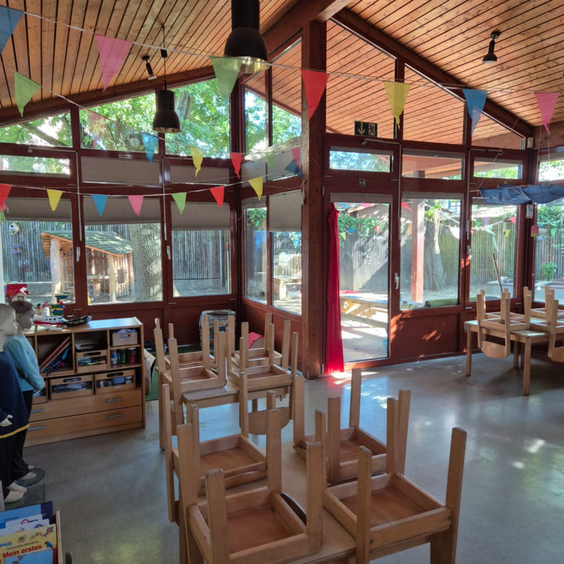Ein sonnendurchfluteter Vorschulklassenraum mit Holztischen, umgedrehten Stühlen und bunten Wimpelketten.