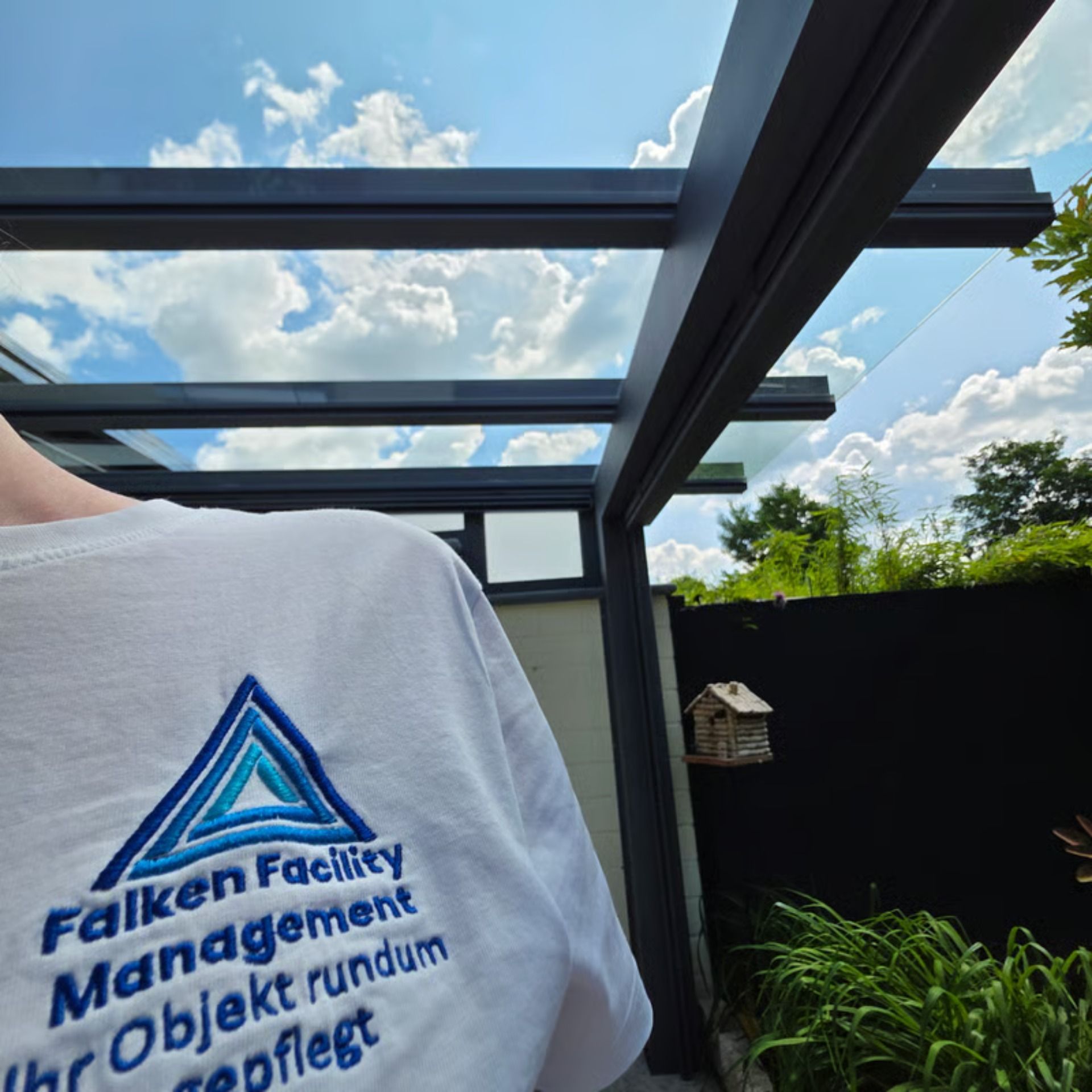 Eine Person mit einem weißen T-Shirt steht in der Nähe einer Glasdachkonstruktion in einem Garten.