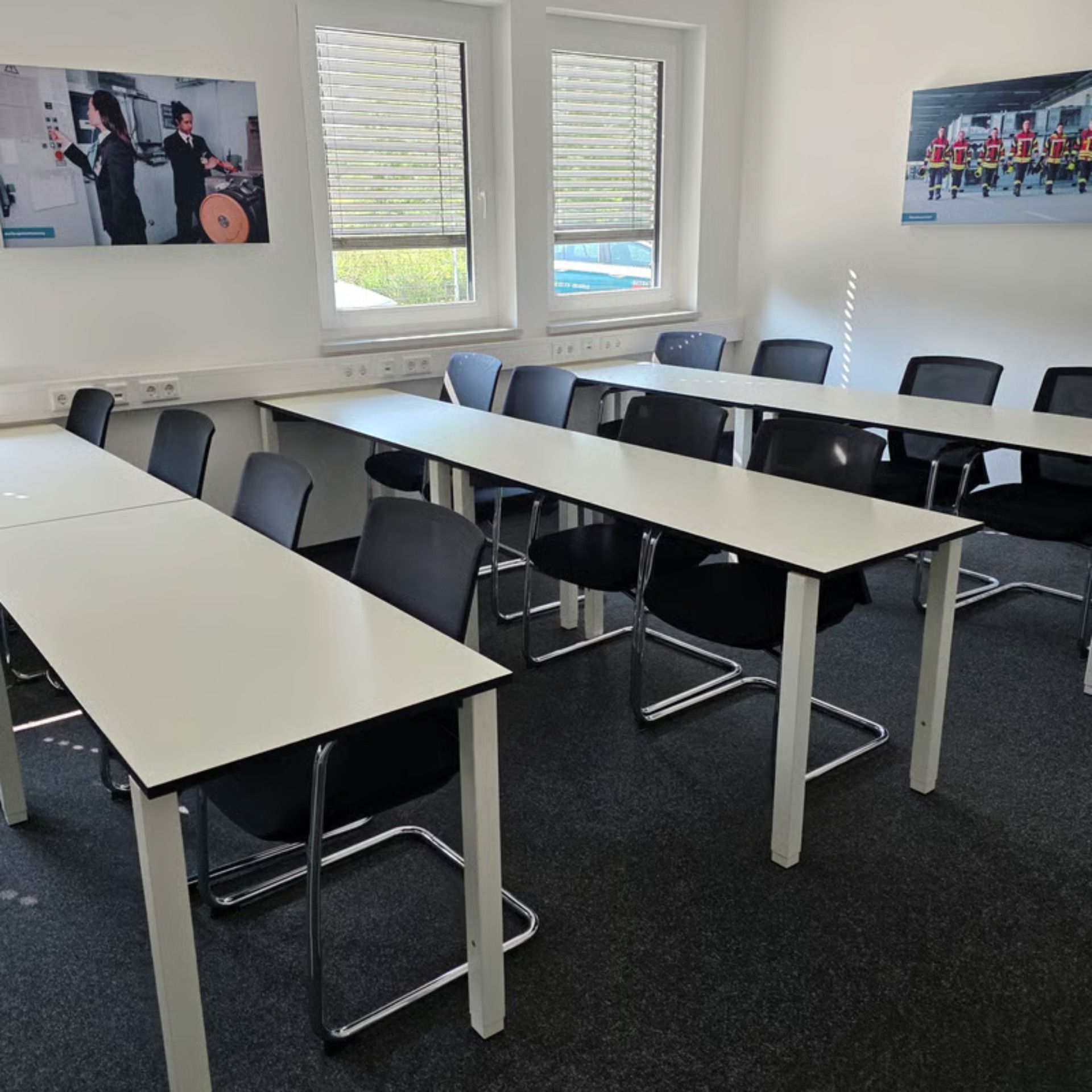 Ein Klassenzimmer mit langen Tischen, schwarzen Stühlen und Fenstern. Zwei Poster sind sichtbar.