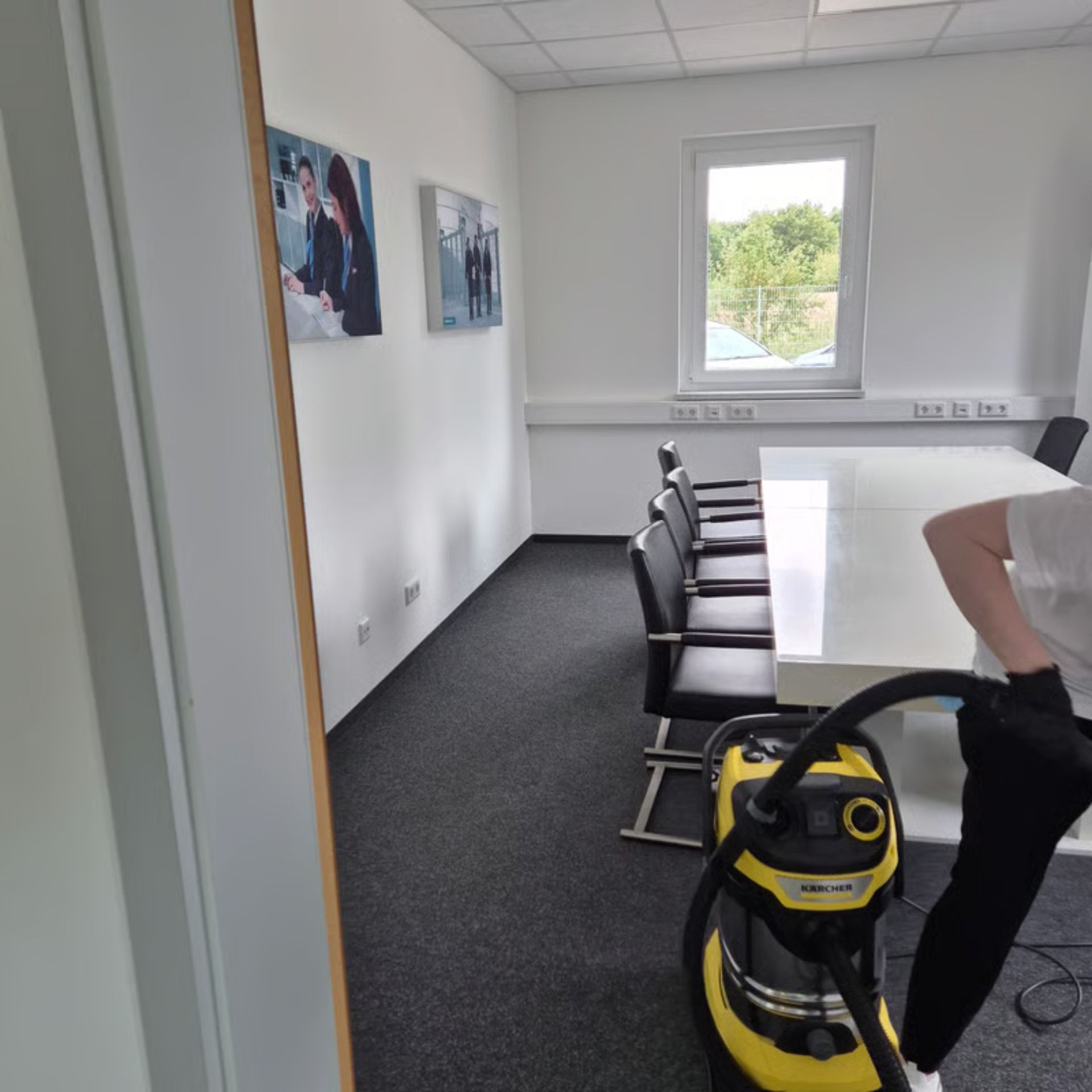 Eine Person saugt einen leeren Konferenzraum mit schwarzem Teppich, weißen Wänden und einem Fenster.