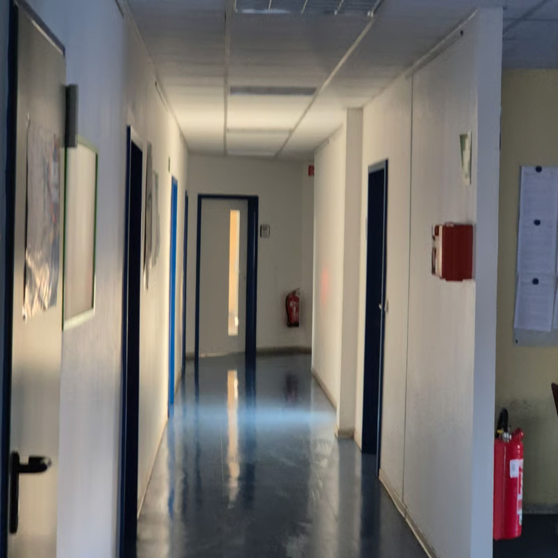 Ein Flur mit weißen Wänden und dunkelblauen Türen. An der Wand hängt ein Feuerlöscher, der Boden glänzt.