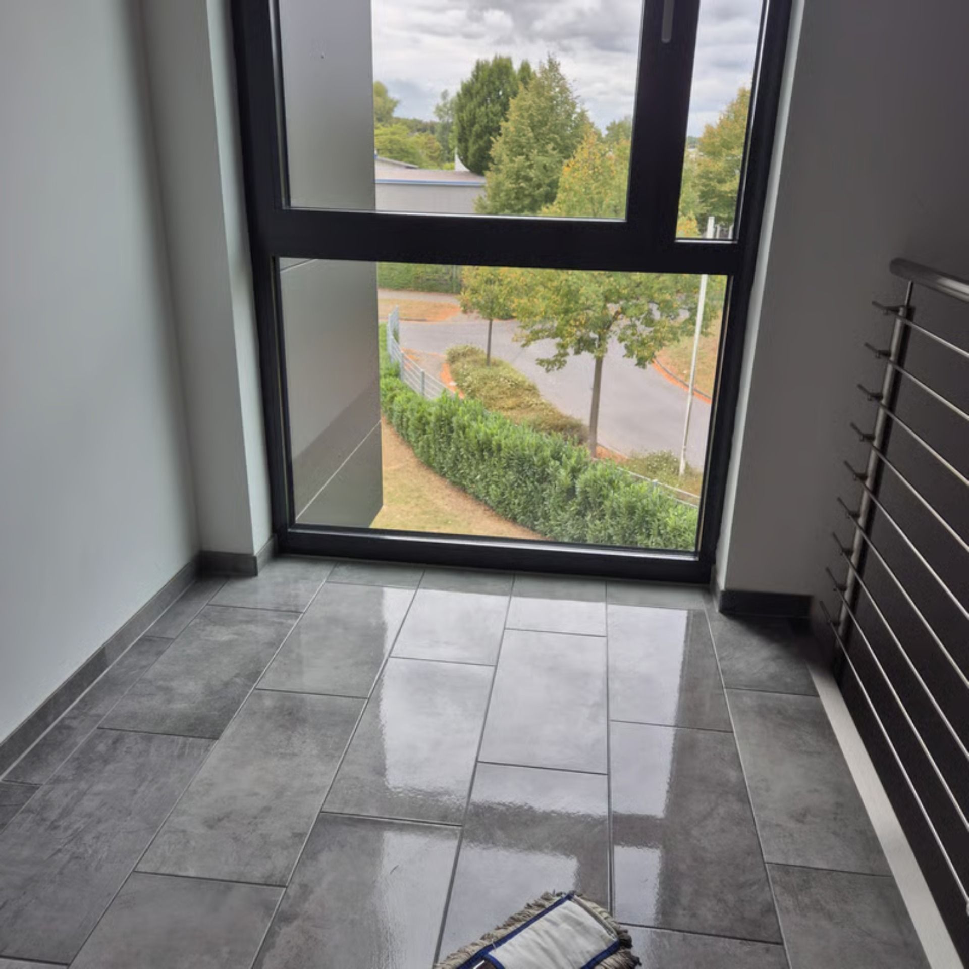 Fenster in einem Flur mit grau gefliestem Boden und Blick auf eine Straße und Bäume unter einem bewölkten Himmel.