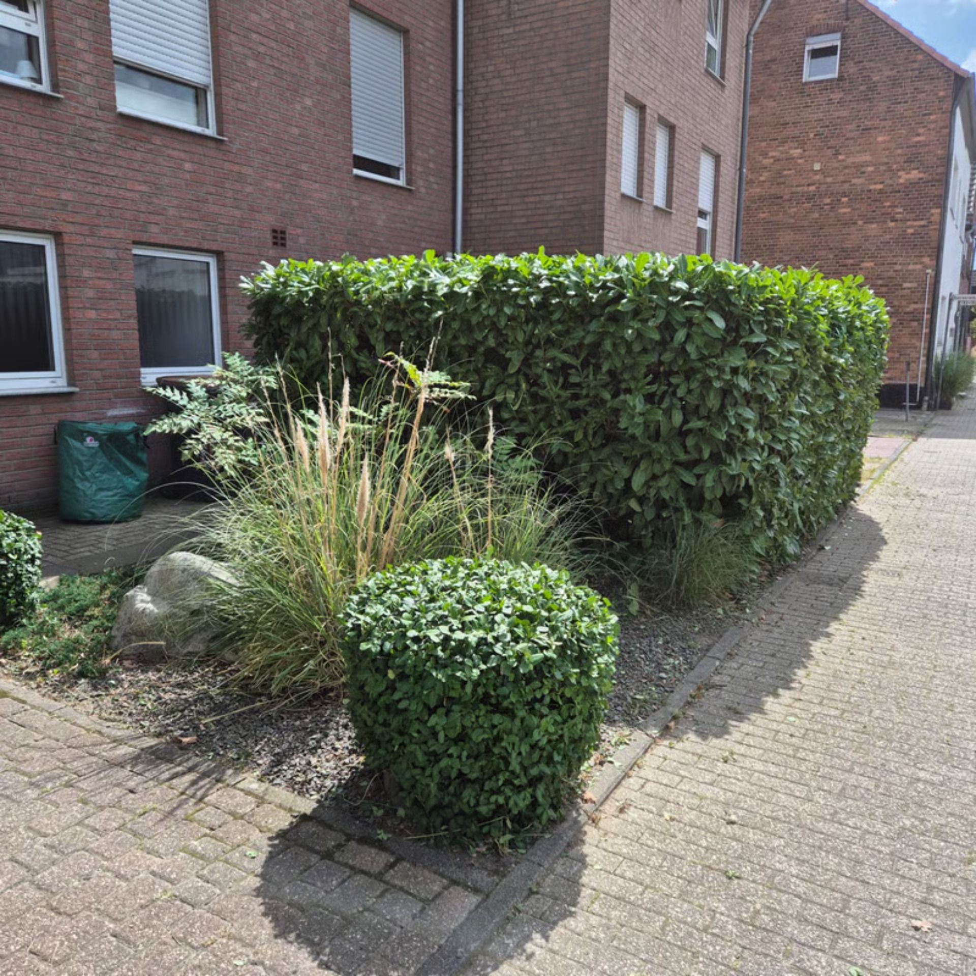 Eine geschnittene grüne Hecke und Sträucher säumen ein Backsteingebäude und einen Backsteinweg.