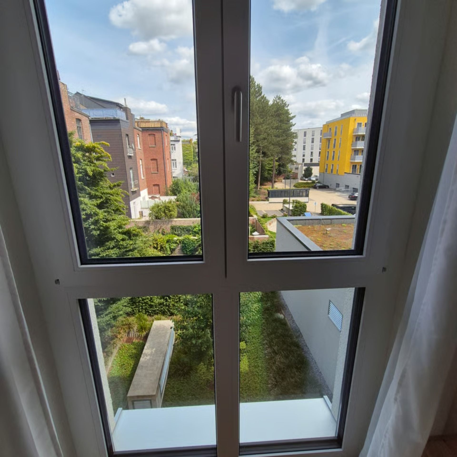 Blick durch ein weißes Fenster: Gebäude, Grünflächen und ein leuchtend gelbes Gebäude unter einem teilweise bewölkten Himmel.