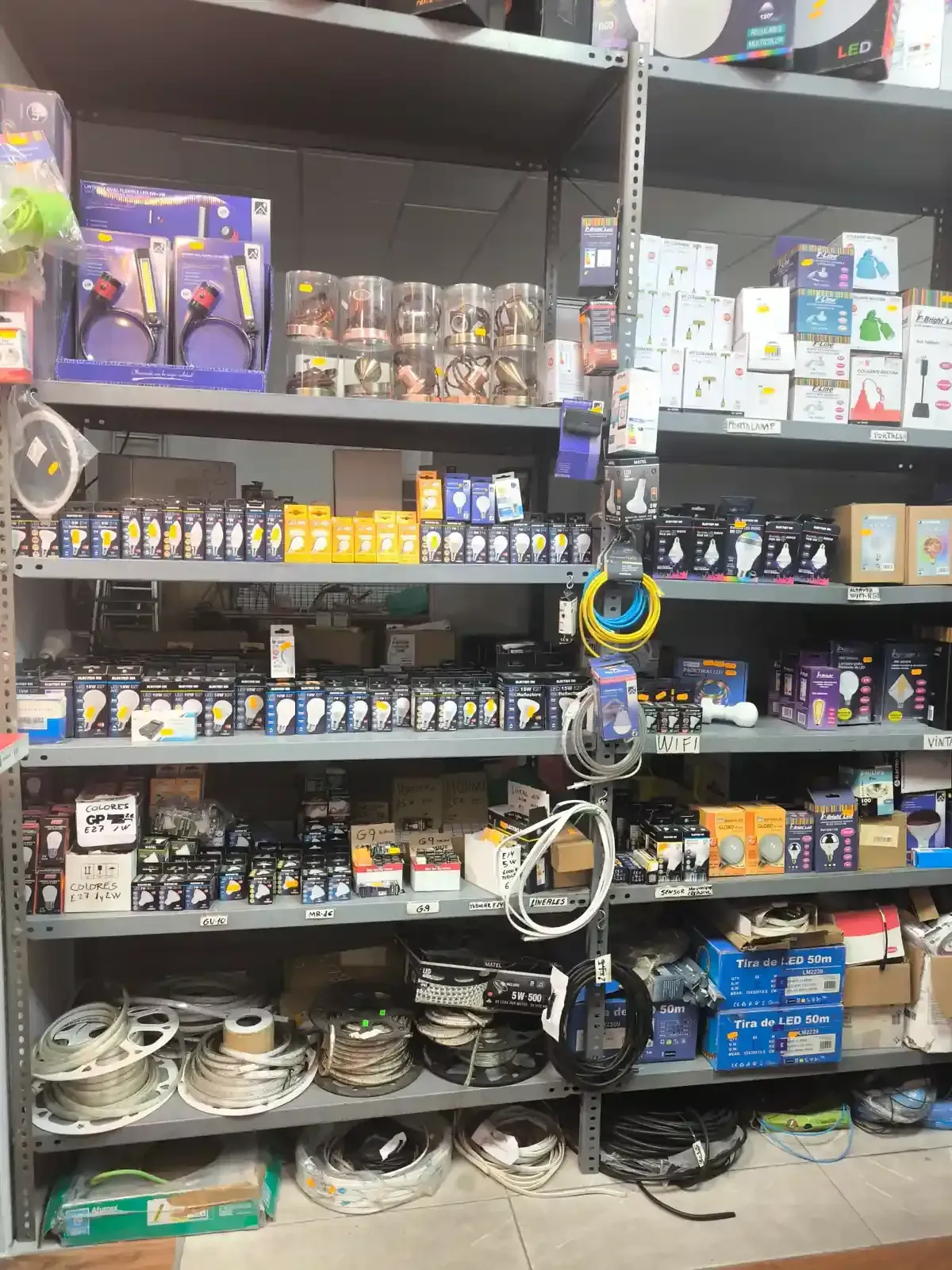 Estantes llenos de componentes eléctricos, incluidas bombillas, cableado y diversos artículos de ferretería en una tienda.