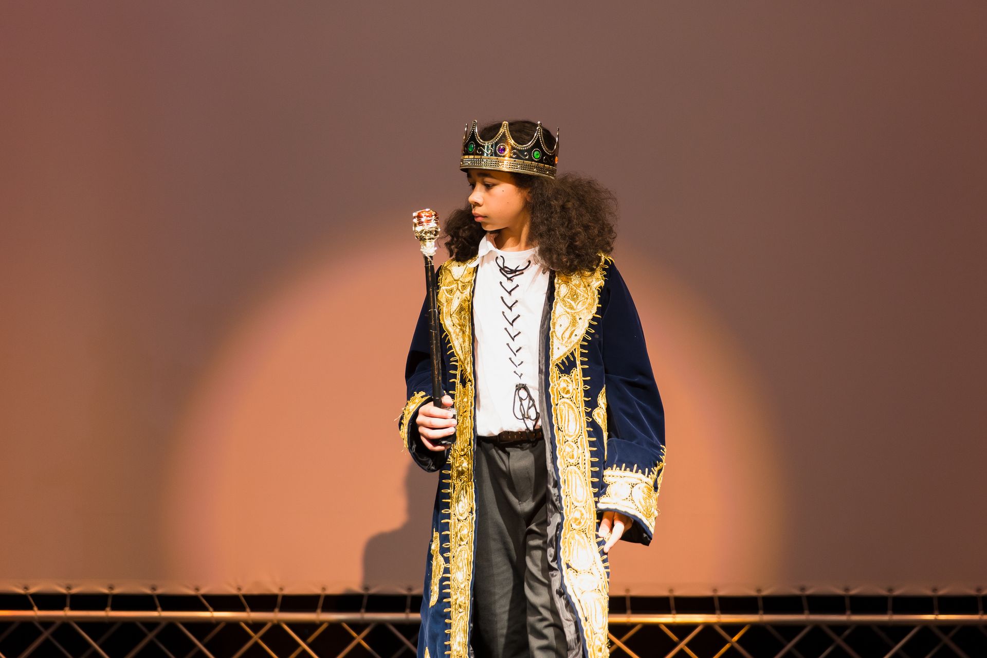 Ein kleiner Junge trägt eine Krone und hält einen Stock auf einer Bühne.
