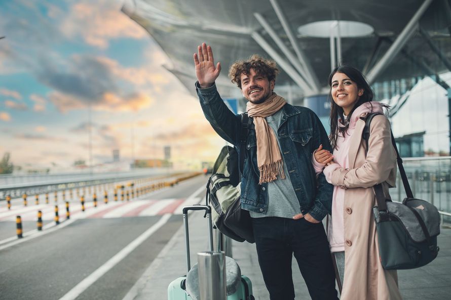 Un couple souriant se tient dans une zone de dépose-minute de l'aéroport avec ses bagages, saluant quelqu'un de la main alors qu'ils se préparent pour un voyage.