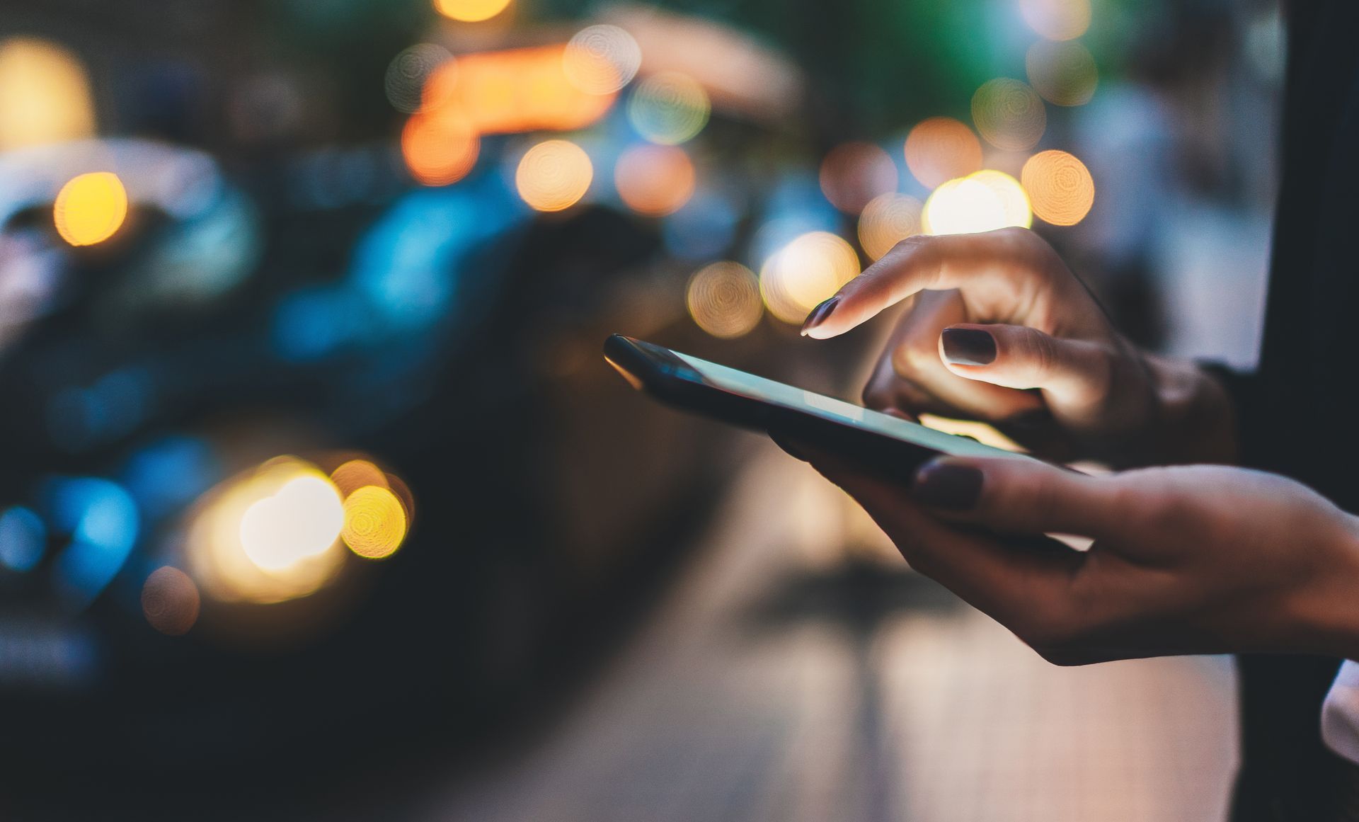 Gros plan sur les mains d'une personne utilisant un smartphone en extérieur, de nuit, avec des lumières de la ville floues en arrière-plan.